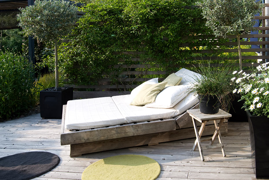 Graue Holzterrasse mit bequemer Gartenliege, dekorativen Kissen und grüner Umrandung aus Sträuchern und Kübelpflanzen.